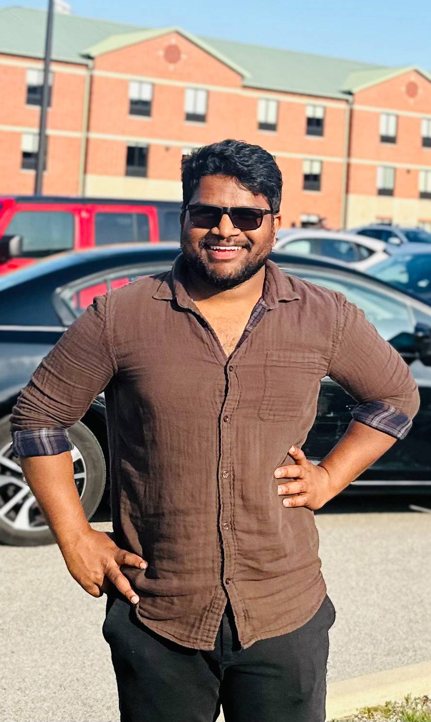 Student in brown shirt and sunglasses smiling at the camera with brick building in the background